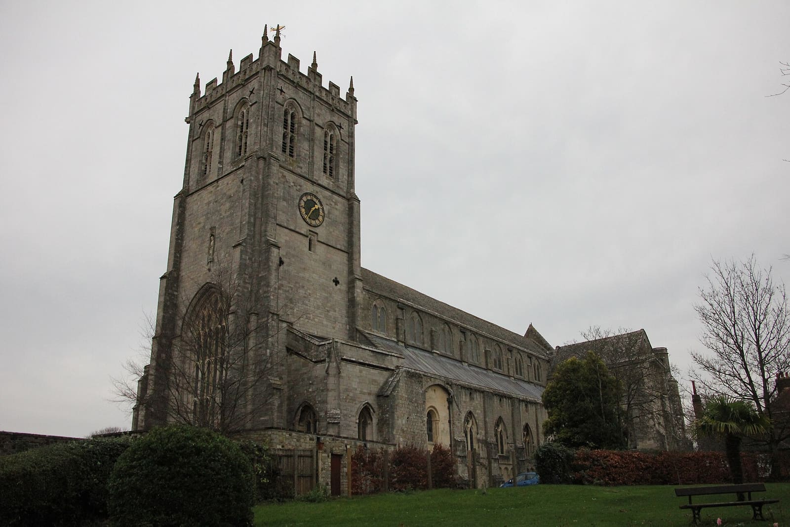 The battlemented tower and long nave of Christchurch Priory rising above its churchyard under a grey sky